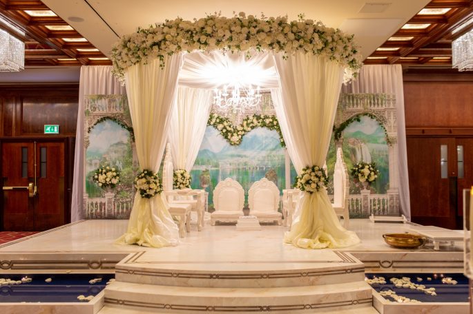 Elegant wedding stage with floral decorations and white chairs under a chandelier.