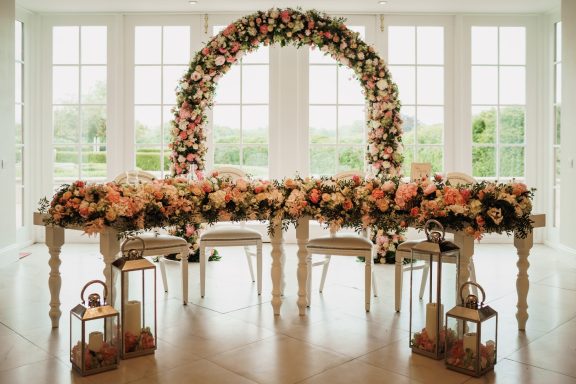 Floral arch above a decorated table with flowers and lanterns in a bright setting.