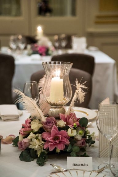 Candle in a glass holder atop floral centrepiece on a dining table.