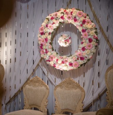 Floral wreath with pink flowers on a decorated wall behind ornate chairs.