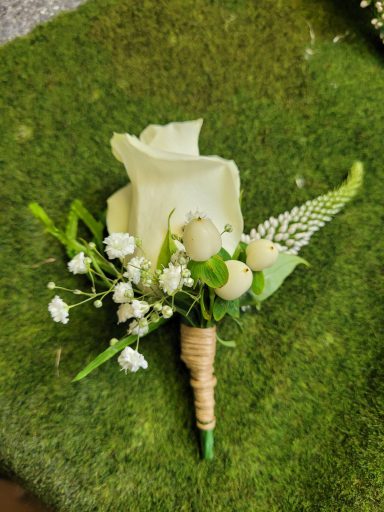 White rose boutonnière with greenery and small white flowers on a green background.