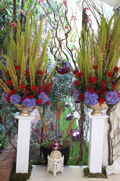 Two large floral arrangements with purple and red flowers in elegant vases on white pedestals.