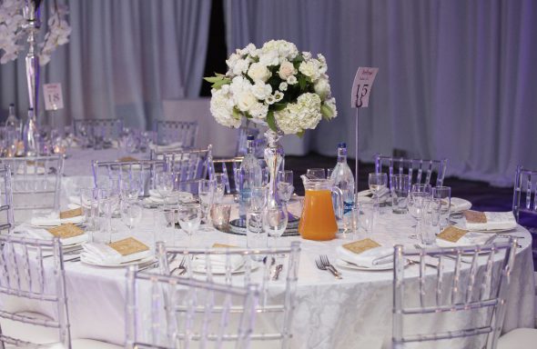 Round dining table set elegantly with white cloth, floral centerpiece, and glassware.