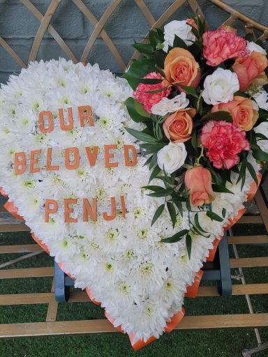 Heart-shaped floral tribute with orange letters spelling "Our Beloved Penji" and roses.
