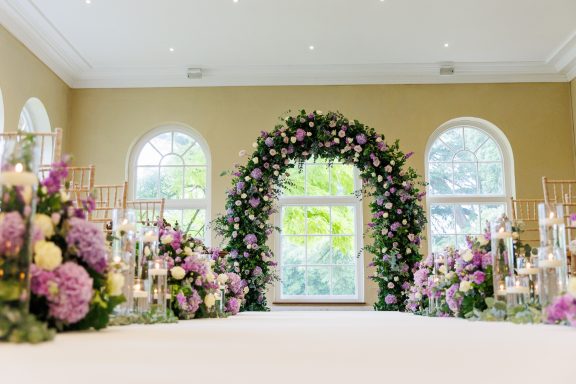 Floral archway adorned with purple and white flowers, flanked by arrangements, in a bright room.
