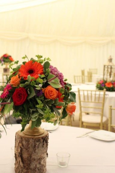 Floral centrepiece with vibrant flowers, set on a rustic wooden base, in a decorated venue.