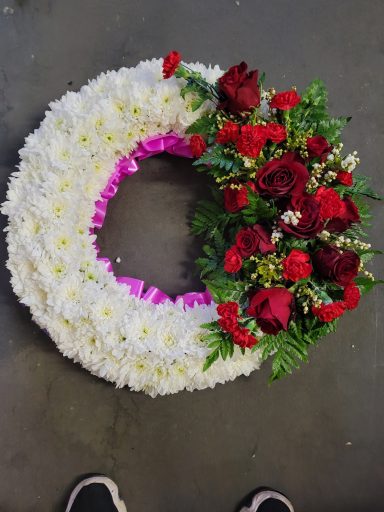 A circular floral wreath with white flowers and red roses, accented with greenery.