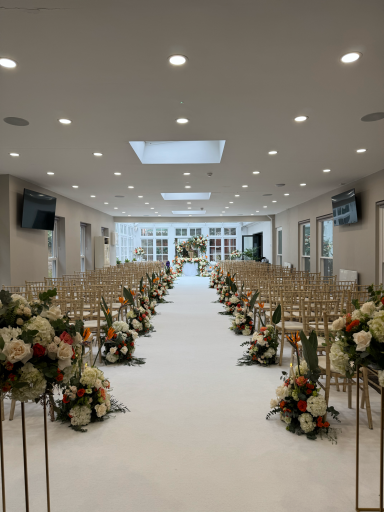 Elegant wedding venue with floral arrangements along a white aisle and golden chairs.